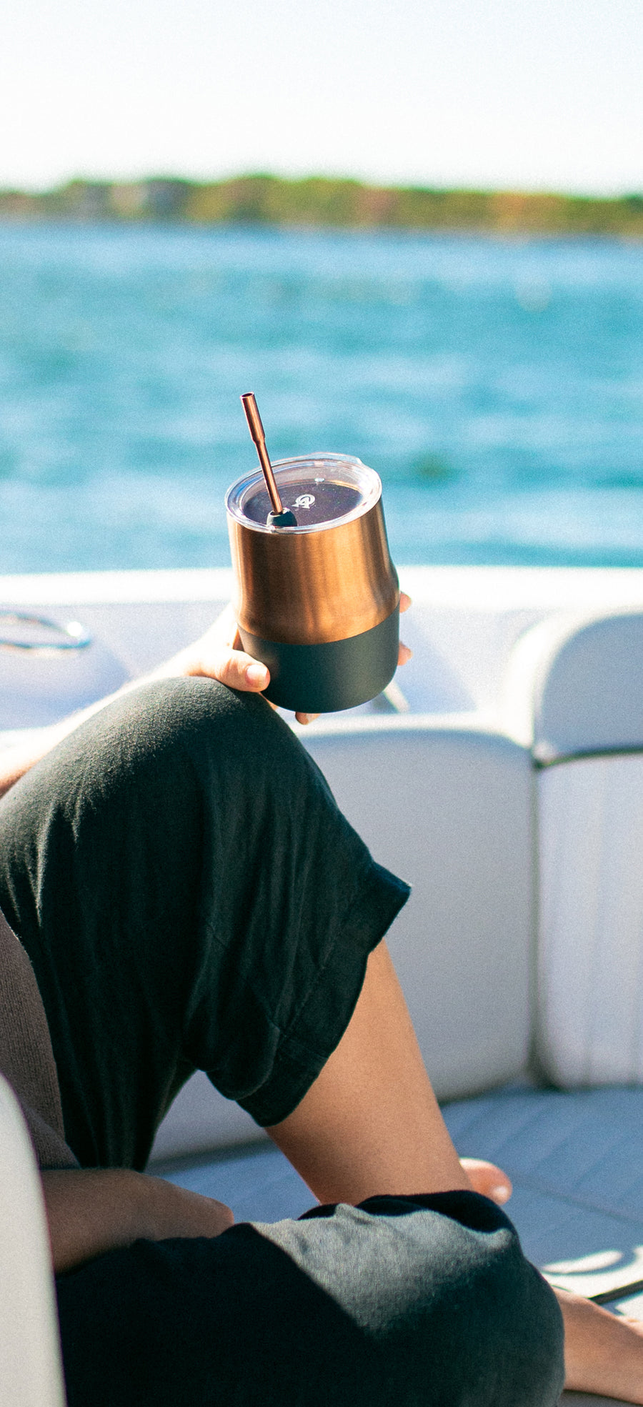 Copper-colored Mule cup with lid and straw held beside blue water