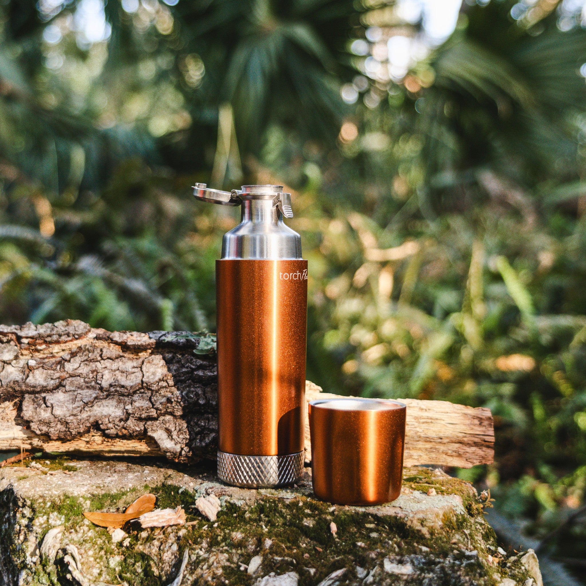 Copper-finish High Camp Torch Flask with its flip lid open and sipping cup alongside on a log